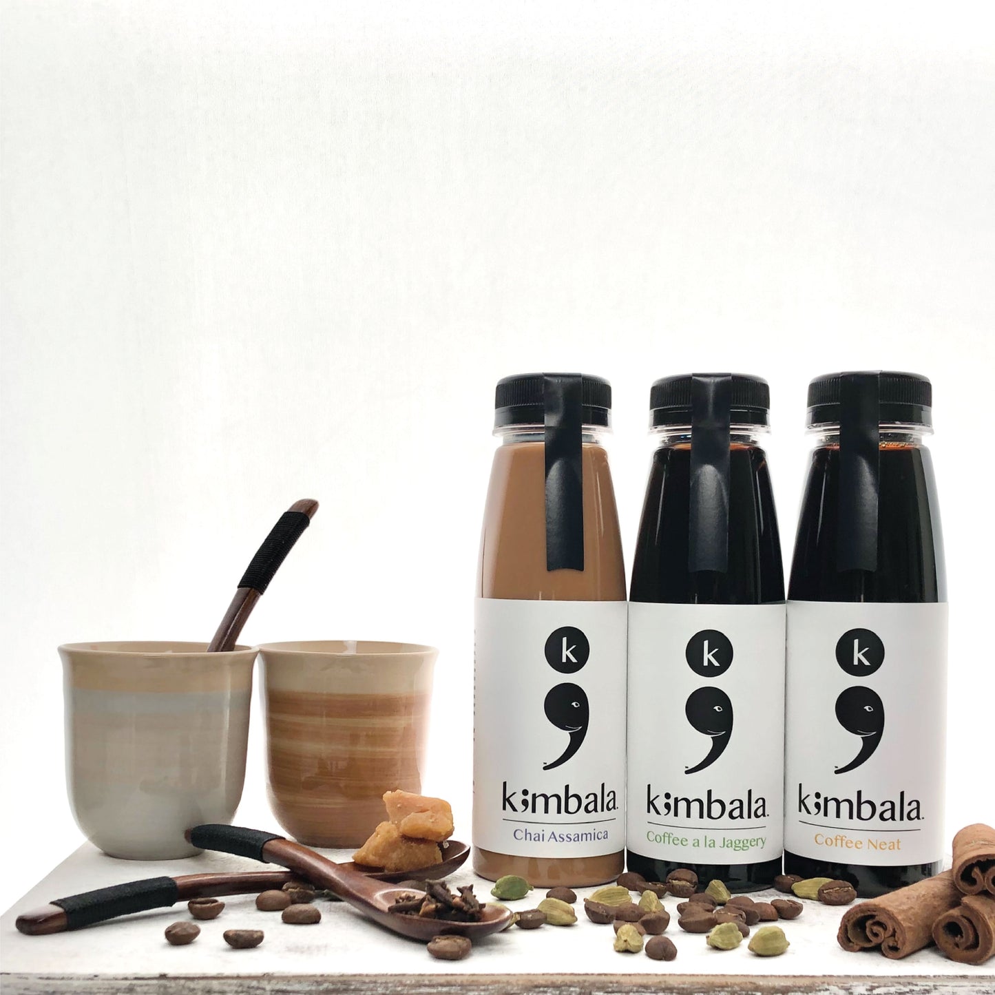 Kimbala chai assamica (10.2oz bottle), Kimbala coffee a la jaggery (10.2oz bottle), Kimbala coffee neat (10.2oz bottle), the best indian chai and clean caffeine black coffee all standing on a light surface with white background surrounded by whole cinnamon sticks, small ceramic cups with blue and brown painted on the side, cardamom pods, cloves, coffee beans and whole pieces of jaggery on small wooden spoons. All products pictured included in the combo pack.