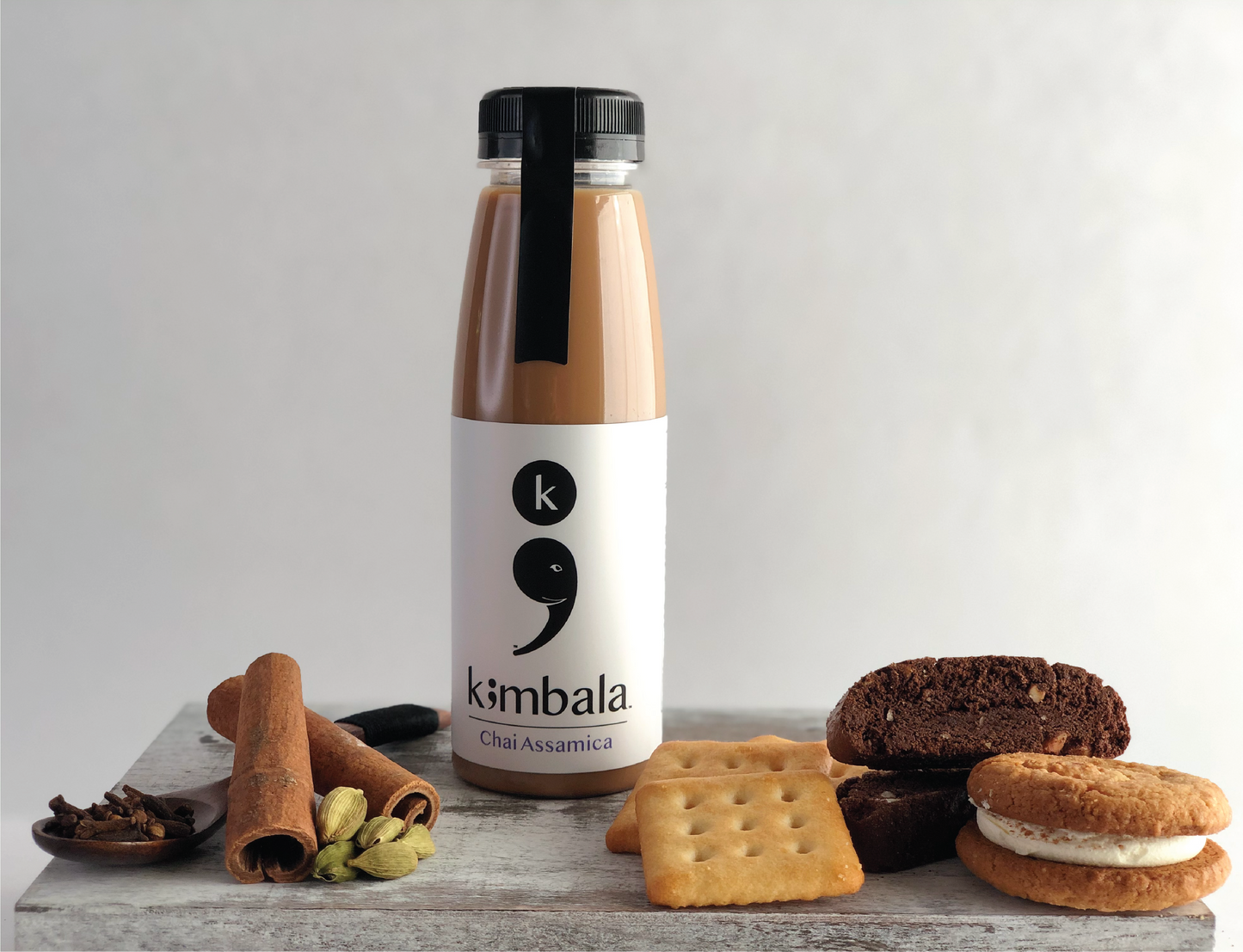 Kimbala Chai Assamica, 10.2oz bottle, the best indian chai surrounded by crackers, biscotti, a cream filled cookie, cinnamon sticks, cardamom pods and cloves on a small wooden spoon