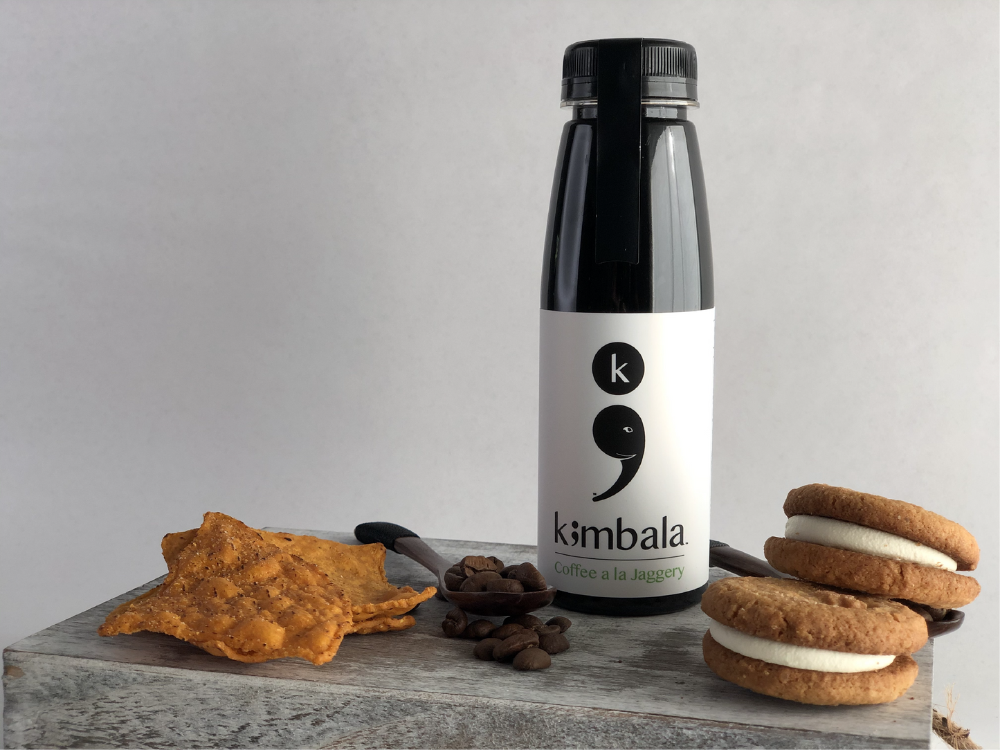 Kimbala Coffee a la jaggery, 10.2oz bottle of clean caffeine black coffee lightly sweetened, surrounded by coffee beans on a small wooden spoon, cream filled cookies and sweet potato chips. Jaggery is an unrefined sugar made from solidified sugar cane juice.