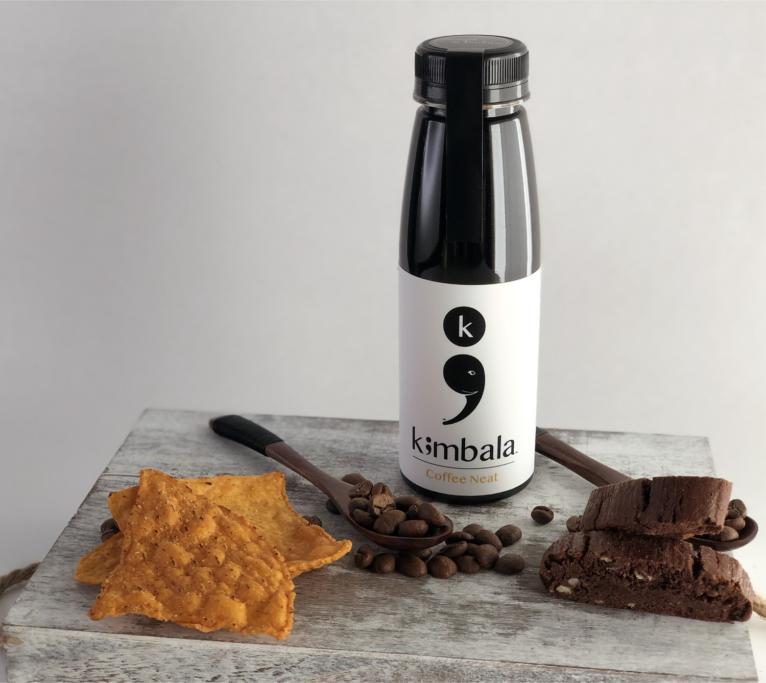 Kimbala Coffee neat, 10.2oz bottle of clean caffeine black coffee, surrounded by coffee beans on a small wooden spoon, chocolate biscotti cookies and sweet potato chips.